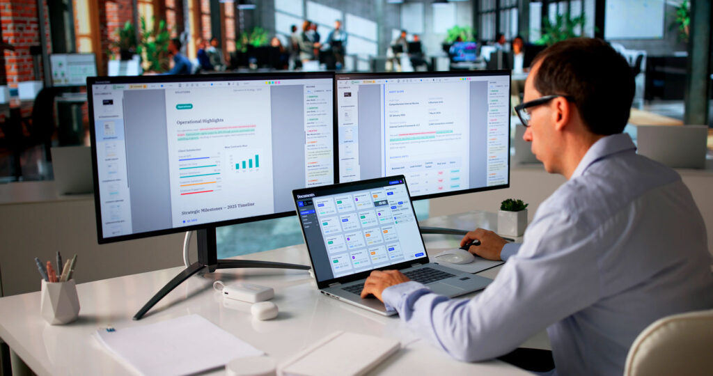IT professional reviewing operational reports on dual monitors and laptop at office desk