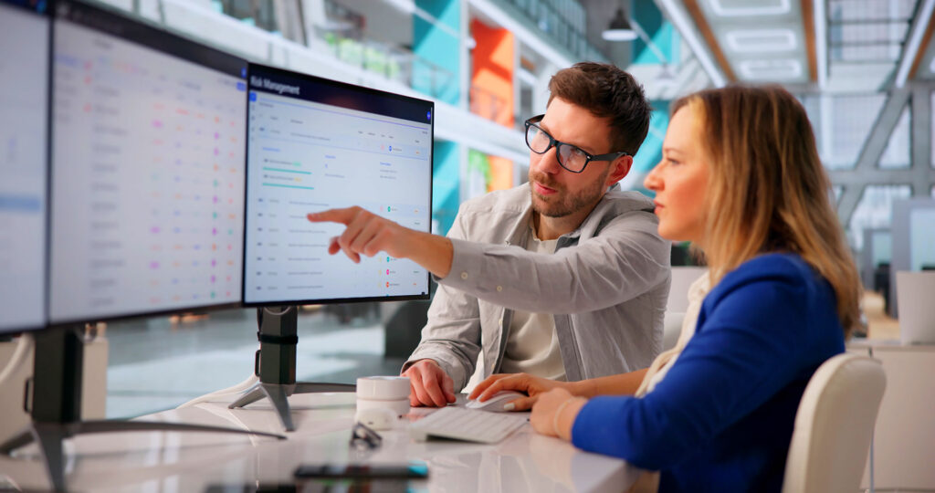 A male IT professional wearing glasses pointing at a computer screen to explain risk management data to a female colleague in a bright office.